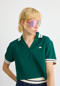 Grønn kortermet polo-skjorte med V-hals, hvite og marineblå stripete mansjetter og liten logo-detalj. Kombinert med store rosa solbriller.