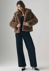 Brun faux fur jakke med en bred krave, knaplukning og en boxy silhuet, parret med højwaistede sorte bukser og spidse hæle.