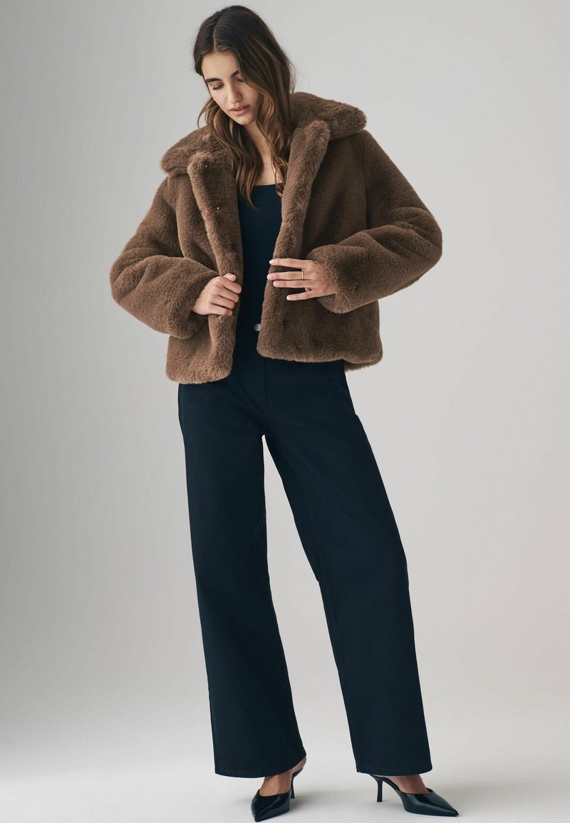 Brun faux fur jakke med en bred krave, knaplukning og en boxy silhuet, parret med højwaistede sorte bukser og spidse hæle.