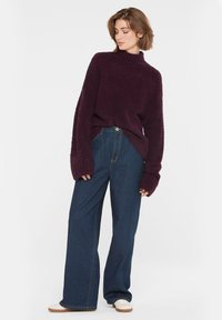 Burgunderroter, flauschiger Pullover mit hohem Kragen und langen Ärmeln, kombiniert mit locker sitzenden, dunklen Jeans und weißen Schnürschuhen.