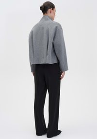 Graue, taillierte Jacke mit strukturiertem Design, weiten Ärmeln und hohem Kragen. Kombiniert mit schwarzen, weit geschnittenen Hosen. Strukturierter Stoff sichtbar.