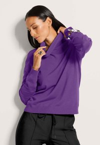 Lila gestrickter Pullover mit hohem umgeschlagenem Kragen, gerippten Bündchen und lässiger Passform. Getragen mit schwarzen Hosen; glatte Textur.