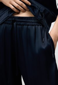 Hand justerar mörkblå satinbyxor med elastisk midja och dragsko, vilket visar en liten del av bar mage.