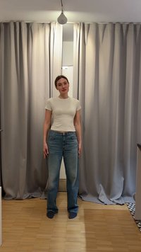 Kvinde med lys hud og brunt hår står foran beige gardiner iført en hvid tætsiddende t-shirt og løse blå jeans på trægulv.