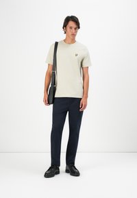Lys beige t-shirt med et lille gult logo, parret med mørkeblå bukser og sorte sko, accessorized med en sort skuldertaske.