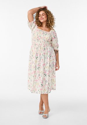 Krøllet kvinde i hvid blomster midi kjole med pufærmer, smilende med den ene arm løftet, iført lyseblå hælede sandaler.