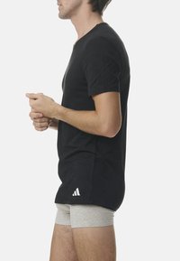 T-shirt nera in cotone con scollo a V, maniche corte e logo a tre strisce bianche vicino all'orlo. Pantaloni corti grigi sono parzialmente visibili sotto.