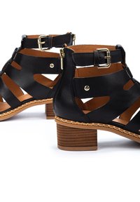 Botas de tobillo de cuero negro con diseño de cortes, tacón bajo, acentos de hardware dorado y detalles trenzados a lo largo de la suela.