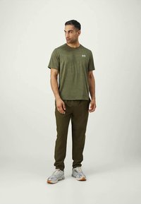 Homem em t-shirt e calças desportivas verde-oliva, olhando de lado, com sapatilhas prateadas e brancas, de pé contra um fundo claro e simples.