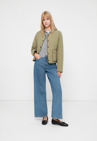 Kurtka pikowana w oliwkowym kolorze z czarnymi zapięciami, szara dzianinowa bluzka i szerokie niebieskie dżinsy. Modelka nosi czarne baleriny, stojąc na białym tle.