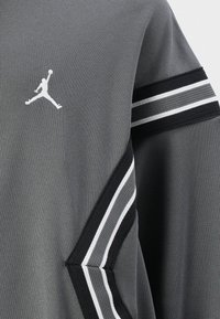 Haut de sport gris en tissu texturé, avec un logo Jumpman blanc et des accents rayés noir et blanc sur les manches.