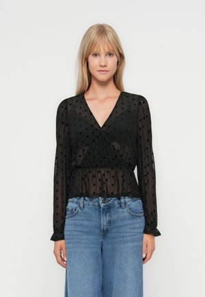Zwarte doorschijnende blouse met stippenpatroon, v-hals, lange mouwen en een peplum taille. Gecombineerd met high-waisted blauwe denimjeans.