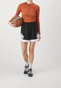 Idrottskläder bestående av en rostfärgad långärmad tröja, svarta shorts med vita detaljer och svarta basket skor. Håller i en basketboll.