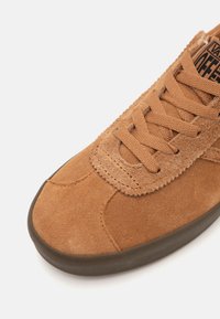 Sapatilha de camurça castanha com parte superior texturizada, atacadores planos e sola de borracha. Possui detalhes cosidos e um design clássico de cano baixo.