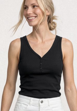 Smilende kvinde med lyst hår i en lav knold har en sort ærmeløs henley-top og hvide bukser på, stående foran en ensfarvet baggrund.