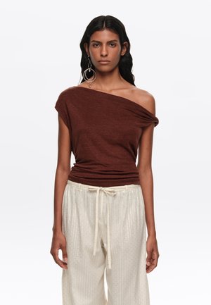 Mujer con un top marrón de un solo hombro, grandes pendientes de aro y pantalones blancos sueltos con cintura ajustable, de pie frente a un fondo liso.