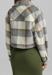 Kobieta ubrana w krótki kraciasty żakiet w odcieniach szarości, bieli i brązu, nałożony na prążkowaną oliwkowozieloną spódnicę, widziana od tyłu.