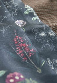 Blumendesign auf Stoff mit dunklem Hintergrund, mit roten, lila und weißen Blumen, neben einem strukturierten braunen Stoff mit einem Rautenmuster.