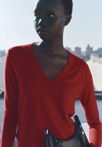 Roter V-Ausschnitt-Pullover aus weichem Strickstoff, mit langen Ärmeln und figurbetonter Form. Kombiniert mit einer schwarzen Handtasche mit metallischen Akzenten.