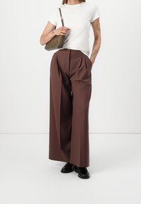 Bruine wijde broek met voorplooien, gecombineerd met een getailleerd wit t-shirt en een kleine olijfgroene tas. Zwarte schoenen maken de look af.