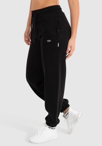 Schwarze, tapering Jogginghosen mit elastischem Bund, Reißverschlusstaschen und gerippten Bündchen. Das Modell trägt weiße Sneaker mit schwarzen Akzenten.