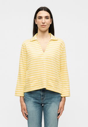 Γυναίκα με ίσια σκούρα μαλλιά φοράει ένα κίτρινο και λευκό ριγέ πουκάμισο με μακριά μανίκια και γιακά και μπλε τζιν, στέκεται μπροστά σε απλό φόντο.