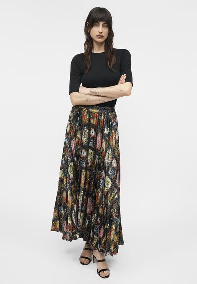 Kobieta o długich ciemnych włosach, ubrana w czarną bluzkę z krótkim rękawem, kolorową plisowaną spódnicę maxi w wzory oraz czarne sandały na obcasie, stoi z założonymi rękami.
