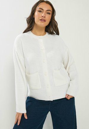 Kvinde iført en hvid knappet cardigan med to forlommer, kombineret med mørkeblå jeans, stående mod en ensartet lys baggrund.