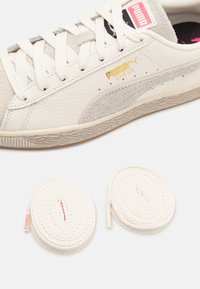 Tenis Puma suede berwarna off-white dengan bagian atas kulit bertekstur, aksen abu-abu muda, dan tali sepatu putih yang dijalin di bawah. Detail logo berwarna emas.
