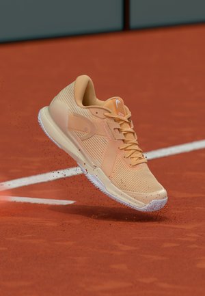 Perzikkleurige sportschoen met een getextureerde mesh bovenkant, een witte zool en een lichtgewicht ontwerp, afgebeeld in de lucht op een gravelbaan.