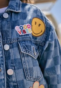 Jeansjacke mit blauem Karomuster, silbernen Knöpfen und bunten gestickten Patches: "Good Vibes" und ein lächelndes Gesicht.
