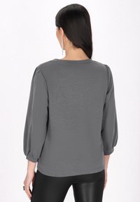 Szürke pulóver, háromnegyedes ujjal, texturált anyagból, kerek nyakkivágással és laza szabással. Fekete leggings-szel viselve.