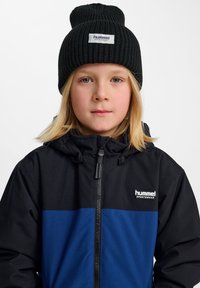 Chaqueta negra y azul con cremallera y capucha, que cuenta con un logo en el pecho. Gorro de punto negro con una etiqueta en la parte delantera.