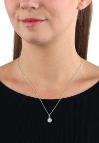 Silverhalsband med en rund berlock som har en blommönsterdesign. Berlocken har flera små stenar, fästa på ett tunt kedja. Bärs över en svart topp.