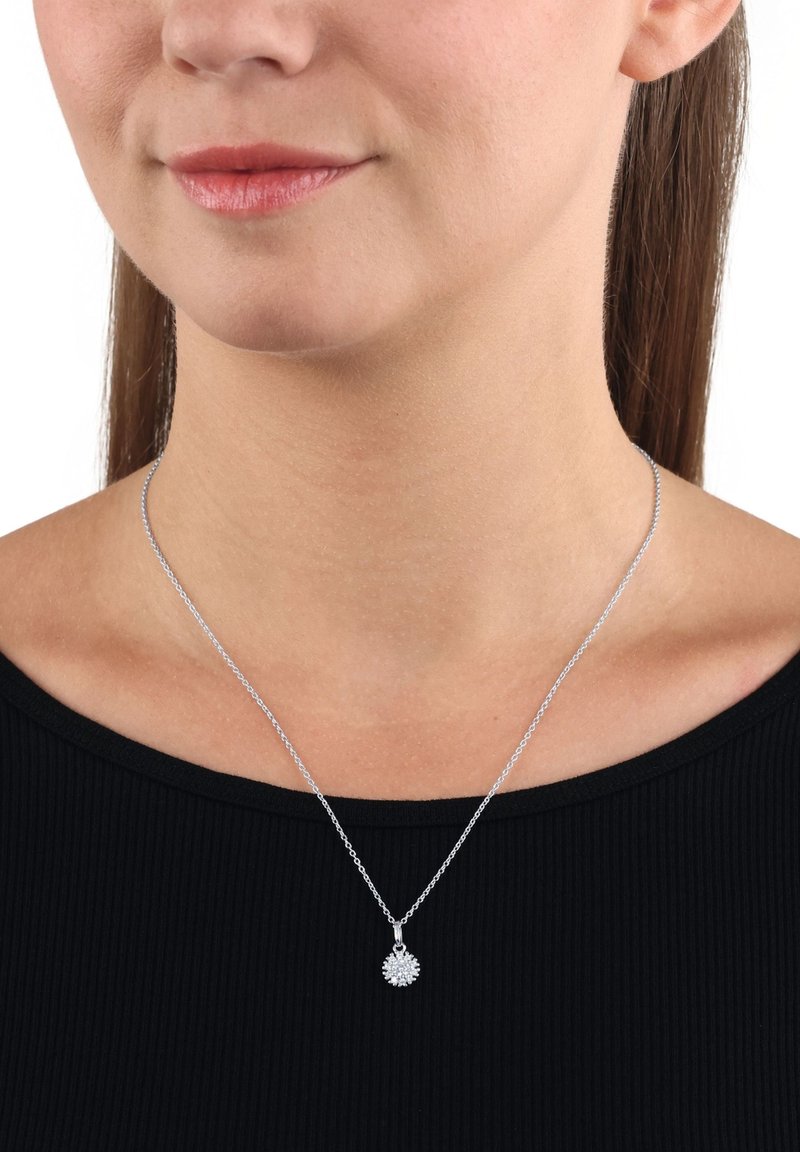 Silverhalsband med en rund berlock som har en blommönsterdesign. Berlocken har flera små stenar, fästa på ett tunt kedja. Bärs över en svart topp.