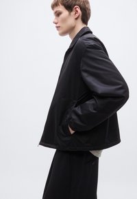 Μαύρο ελαφρύ μπουφάν με υφή στην επιφάνεια, άνετη εφαρμογή και ελαστικά μανίκια, φορεμένο πάνω από ανοιχτόχρωμο πουκάμισο και μαύρο παντελόνι.