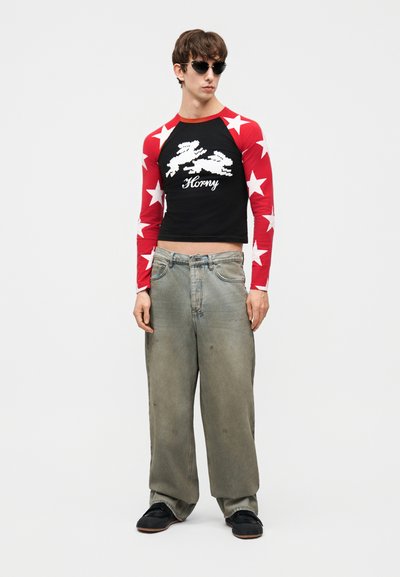 Young man wearing pixelated bunny black crop top with red star-patterned sleeves, loose gray jeans, black shoes, and sunglasses, standing.