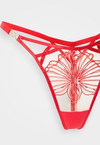 Εσώρουχο στρινγκ από κόκκινη δαντέλα με περίπλοκη κεντητή λουλουδάτη διακόσμηση σε διαφανές ύφασμα, με ρυθμιζόμενους ράβδους και λεπτομέρειες σε χρυσό τόνο.