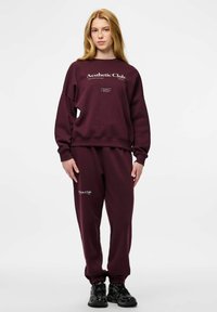 Młoda osoba stojąca w bordowej bluzie i spodniach dresowych z napisem "Aesthetic Club", ubrana w czarne trampki na białym tle.