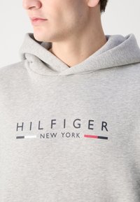 Jeune personne portant un sweat à capuche gris clair avec le logo "Hilfiger New York" en noir, blanc et rouge sur la poitrine, tournée vers la gauche.