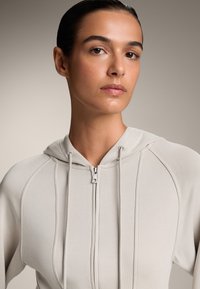 Felpa zip a cappuccio beige chiaro con un design aderente, dotata di cappuccio, lacci, texture a costine e dettagli di cucitura.