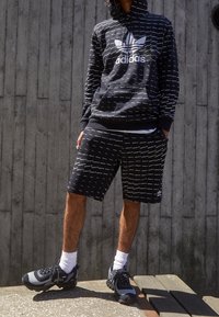 Svart hoodie och shorts med ett vitt upprepande textmönster som har Adidas-logotypen. Svarta och grå sneakers, vita strumpor. Avslappnad atletisk stil.