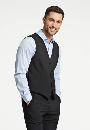 Zwarte formele vest over een lichtblauw overhemd, met knopen aan de voorkant, een zachte textuur en twee zijzakken. Donkere broek is gedeeltelijk zichtbaar.