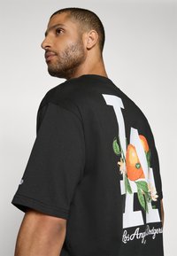 Sort bomulds T-shirt med et stort hvidt "LA" design på bagsiden med orange frugt og grønne blade, korte ærmer og rund halsudskæring.