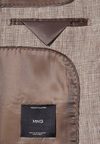 Doublure intérieure d'une veste de costume pied-de-poule marron et beige avec une étiquette noire cousue portant la mention "MNG The Suit Collection, made in India."
