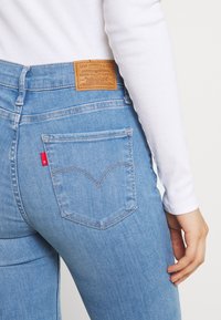 Jeans de mezclilla azul claro con una cintura alta, mostrando un bolsillo trasero con un diseño de arco característico y una etiqueta de cuero marrón en la cintura.