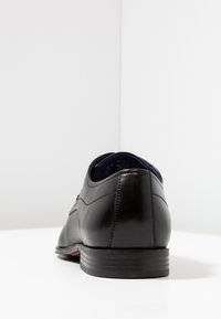Zapato de vestir de cuero negro visto desde atrás, con un acabado suave, punta redondeada y un tacón bajo apilado. El forro parece ser azul.