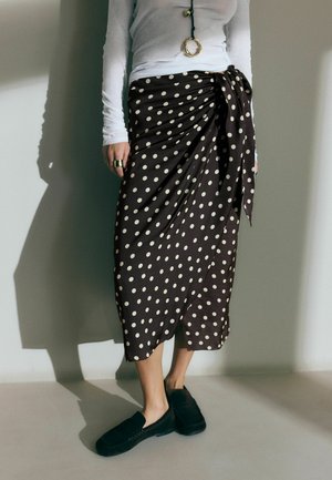 Osoba ubrana w biały top z długim rękawem oraz czarną spódnicę midi w białe kropki, wiązaną w talii, w połączeniu z czarnymi wsuwanymi butami.