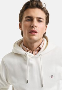White hoodie with drawstrings and silver hardware, layered over a light plaid shirt. Features a small logo on the chest.