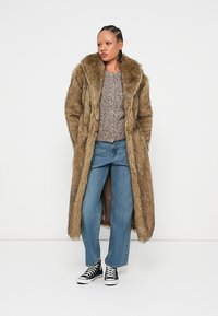 Μακρύ καφέ ψεύτικο γούνα παλτό με φαρδύ γιακά, συνδυασμένο με πλεκτή μπλούζα, μπλε τζιν και μαύρα αθλητικά παπούτσια ψηλής κοπής.